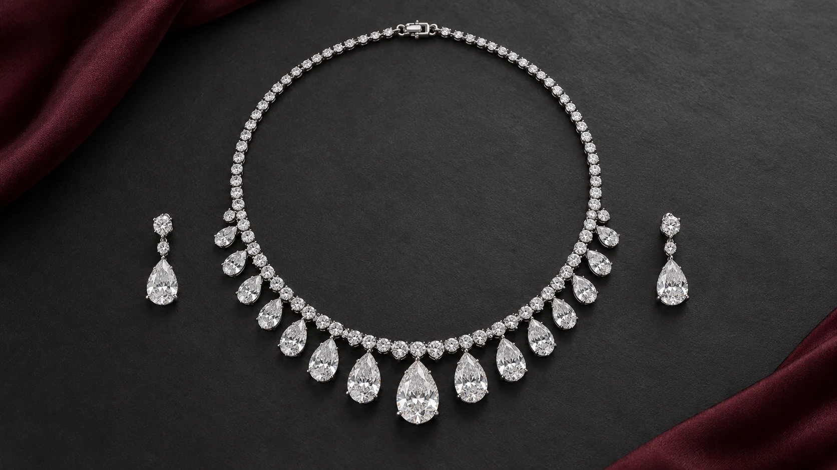 Light held in a setting — complete white-metal clear-stone necklace with pear-shaped drops graduated along a single rounded claw-set line and two matching pear-drop earrings on charcoal
