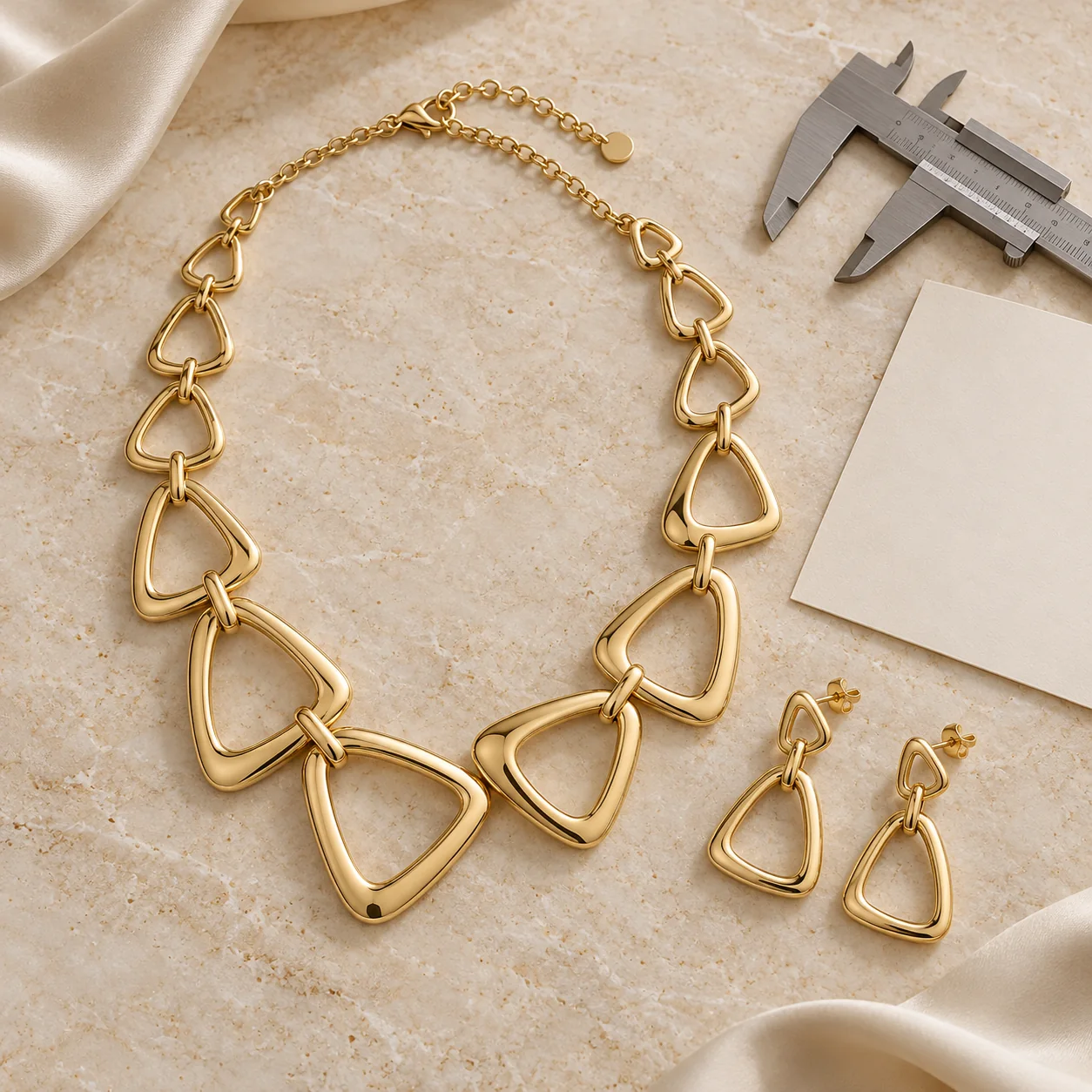Custom Jewellery collection - completed yellow-gold asymmetric necklace of smooth rounded triangular hollow links with two matching open triangular drop earrings beside unbranded calipers and a plain blank cream paper sheet, no writing